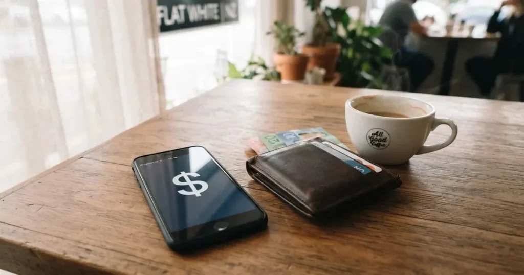 A smartphone on a wooden table beside a wallet and a coffee cup